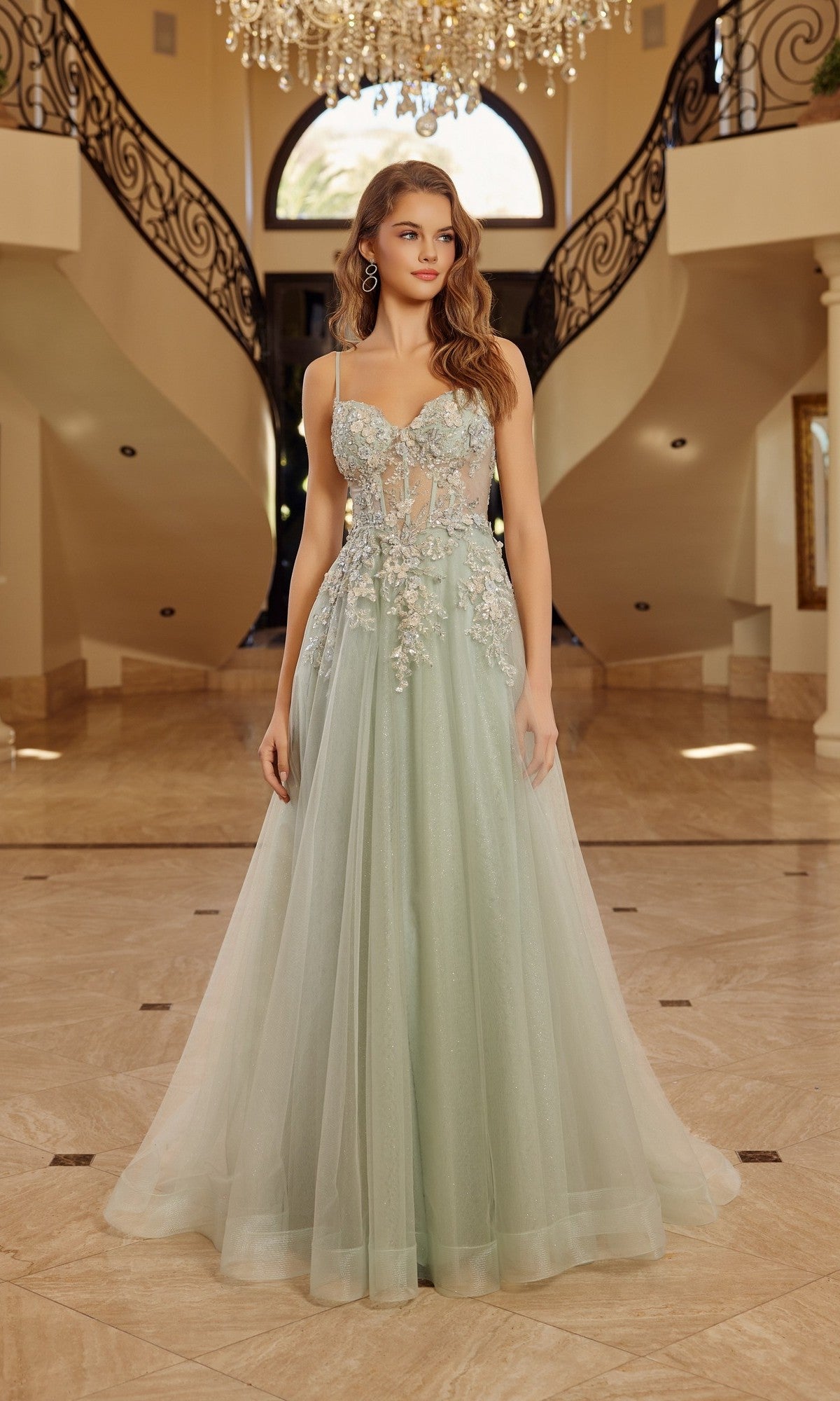 A woman stands in a spacious foyer beneath a chandelier and between two curved staircases, wearing the Nox Anabel G1787 long prom dress in sage green, featuring 3D floral appliques and an elegant A-line silhouette.